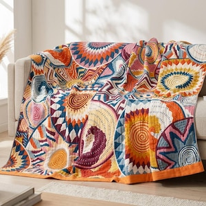 Puede incluir: Una manta acolchada con un patrón multicolor vibrante de círculos concéntricos, espirales y motivos geométricos de rayos de sol en tonos naranja, amarillo, azul, rosa y morado, terminada con un borde de tela de color naranja sólido.