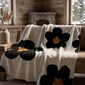 Puede incluir: Una manta de textura color crema con un patrón repetitivo de flores grandes de color negro con centros de color amarillo mostaza. La tela tiene una apariencia suave, difusa y afelpada, con el diseño floral distribuido por toda la superficie.