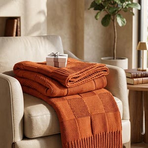 Könnte beinhalten: Eine rostorange Strickdecke mit strukturiertem Schachbrettmuster und gerippten Rändern, die über einen cremefarbenen Sessel drapiert ist. Eine kleine weiße Geschenkbox mit grauem Band liegt auf dem gefalteten Stoff der Decke.