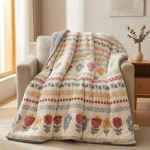 Könnte beinhalten: Eine cremefarbene Wohndecke mit einem sich wiederholenden Muster aus horizontalen Streifen, geometrischen Formen, Punkten und gestickten Blumen in Rot-, Gelb- und Blautönen. Die Decke hat eine weiche, strukturierte Oberfläche und ein graues Fleecefutter.