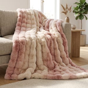 May include: A plush, faux fur throw blanket featuring a textured, tufted design with a gradient color pattern that transitions between soft shades of cream, beige, and dusty rose, draped over the arm and seat of a neutral-toned sofa.