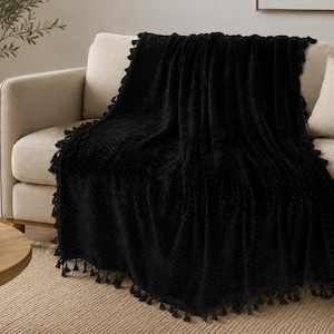 May include: A solid black throw blanket with a textured, ribbed surface and decorative tassels along the edges. The blanket is draped over a light-colored sofa, showcasing its deep black color and the intricate fringe detail on all sides.