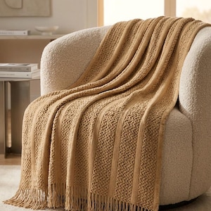 Op de afbeelding: Een mosterdgele gebreide plaid met afwisselende verticale banen van gladde tricotsteek en een gestructureerd mandvlechtpatroon, afgewerkt met franjes aan de onderkant, gedrapeerd over een crèmekleurige fauteuil.