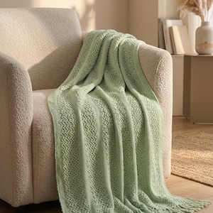 Può includere: Una coperta in maglia color verde salvia chiaro caratterizzata da pannelli verticali testurizzati alternati e motivi traforati, rifinita con un bordo a frange. La coperta è drappeggiata su una poltrona testurizzata color crema.