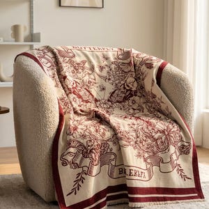 Op de afbeelding: Een crèmekleurige plaid met een donkerrode botanische print en vlinderpatronen. Het ontwerp bevat gedetailleerde bloemmotieven, diverse vlinderillustraties en decoratieve linten met de tekst BUTTERFLY aan de bovenzijde gedrukt.
