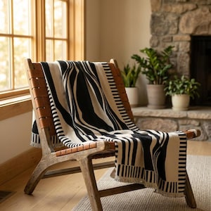 Peut inclure: Un plaid tissé présentant un motif géométrique audacieux noir et crème avec des rayures verticales et des formes abstraites, fini avec un bord à franges. Le plaid est drapé sur une chaise en bois avec des lanières en cuir marron.