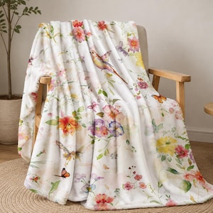 Puede incluir: Una manta de forro polar blanca con un estampado de acuarela de flores coloridas, follaje verde, mariposas y un pájaro pequeño. La tela suave muestra un patrón botánico repetitivo con tonos de flores rojas, amarillas, rosas y moradas.