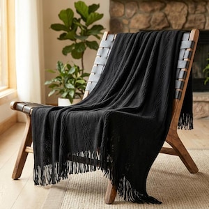 Könnte beinhalten: Eine einfarbige schwarze Strickdecke mit vertikalem Wellenmuster und langen Fransen am unteren Rand. Die Decke ist über einen Holzstuhl mit schwarzen Lederriemen drapiert und zeigt ihre weiche, strukturierte Stoffbeschaffenheit.