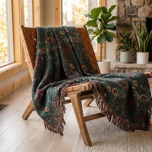 May include: A dark teal woven throw blanket featuring a repeating floral pattern in shades of red, yellow, and green. The blanket has a textured surface and is finished with long, dark brown fringe along the edges, draped over a wooden chair.