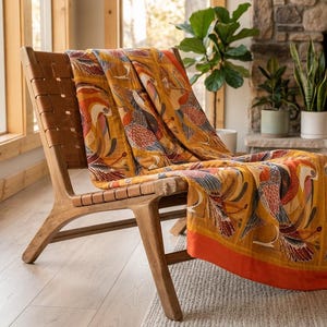 Può includere: Una sedia in legno con seduta e schienale intrecciati, drappeggiata con una coperta color giallo senape caratterizzata da un motivo ripetuto di uccelli stilizzati nei toni dell'arancione, blu e bianco, rifinita con un bordo arancione.