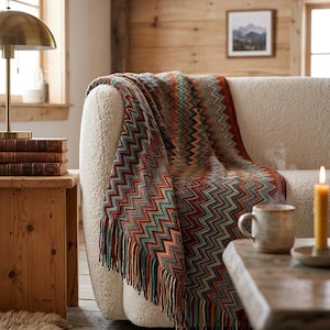 May include: A woven throw blanket featuring a multi-colored chevron pattern in shades of rust, teal, mustard yellow, and cream. The blanket has a textured knit construction and is finished with long, multicolored fringe along the bottom edge.