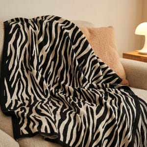 Pode incluir: Uma manta de malha com um padrão de riscas de zebra em preto e creme de alto contraste. O tecido tem uma textura suave e entrelaçada com uma borda preta sólida ao longo das extremidades, drapeada sobre um sofá de cor neutra e texturizada.