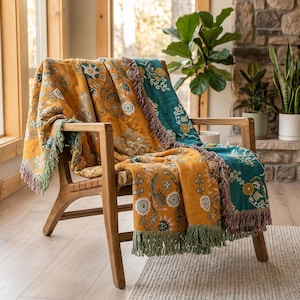 Op de afbeelding: Een geweven plaid gedrapeerd over een houten stoel, met een tweekleurig ontwerp met mosterdgele en groenblauwe secties. De stof toont een herhalend bloemen- en botanisch patroon in wit en goud, afgewerkt met decoratieve franjes aan de randen.