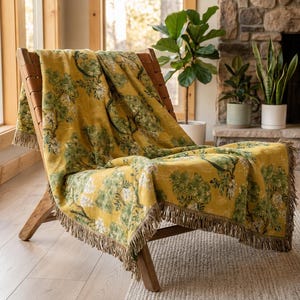 Pode incluir: Uma manta amarelo-mostarda com um padrão repetitivo de árvores verdes e brancas e franjas nas bordas, drapeada sobre uma cadeira de madeira com tiras de couro, exibindo uma construção de tecido texturizado em toda a peça.
