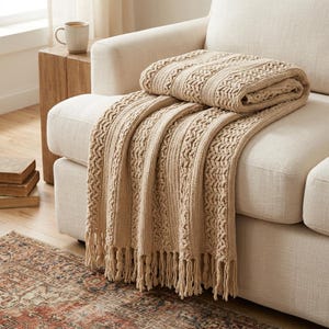 May include: A beige knit throw blanket featuring a textured cable knit pattern with vertical ribbed sections and knotted fringe trim along the bottom edge. The blanket is folded and draped over the arm and seat of a light-coloured upholstered sofa.