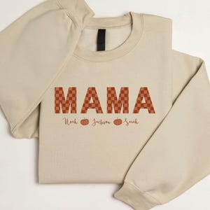 Peut inclure: Un sweat-shirt beige à col rond arborant le mot MAMA dans un motif à carreaux couleur rouille. Sous le texte, les prénoms Noah, Jackson et Sarah sont imprimés dans une police cursive, séparés par de petites illustrations de citrouilles.
