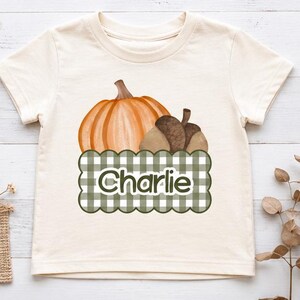 Puede incluir: Una camiseta de manga corta color crema con un gráfico de una calabaza naranja y una bellota marrón sobre un rectángulo festoneado de cuadros vichy verdes y blancos. El nombre Charlie está impreso en letras blancas dentro del rectángulo.