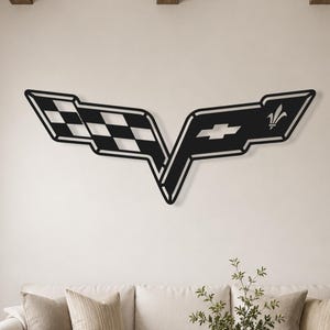 May include: A black metal wall art piece shaped like the Chevrolet Corvette emblem, featuring a chequered flag pattern on the left side and a bowtie logo with a fleur-de-lis symbol on the right side, mounted on a plain off-white wall.
