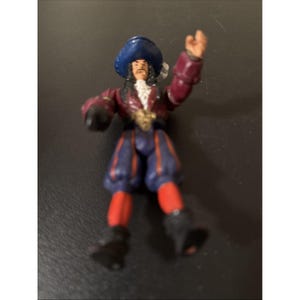 May include: A small plastic action figure of a pirate character wearing a blue hat, a maroon jacket with a white ruffled collar, and blue breeches with red stripes. The figure has long dark hair, a mustache, and one arm raised in a waving gesture.