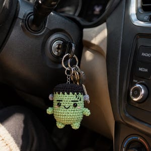 May include: A small, handmade crochet keychain featuring a green, square-headed character with black hair, a stitched mouth, and small grey bolts on the sides of its head. The keychain is attached to a metal ring holding several silver-colored keys.