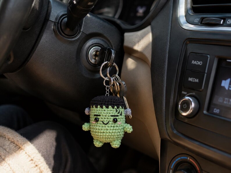 May include: A small, handmade crochet keychain featuring a green, square-headed character with black hair, a stitched mouth, and small grey bolts on the sides of its head. The keychain is attached to a metal ring holding several silver-colored keys.