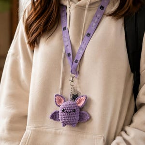 May include: A light purple crochet bat plushie attached to a matching purple lanyard. The bat features pink inner ears, black embroidered eyes, and a small smile. The lanyard is decorated with a repeating pattern of small black cat faces.