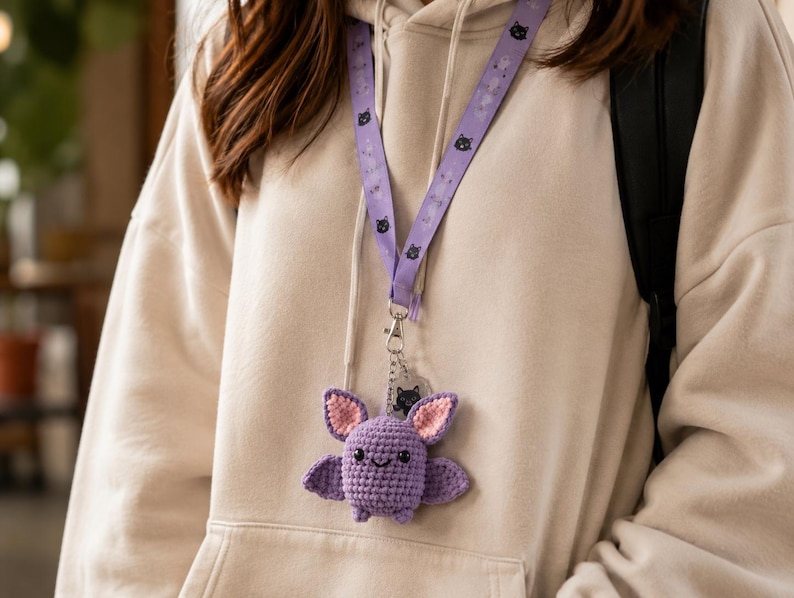 May include: A light purple crochet bat plushie attached to a matching purple lanyard. The bat features pink inner ears, black embroidered eyes, and a small smile. The lanyard is decorated with a repeating pattern of small black cat faces.