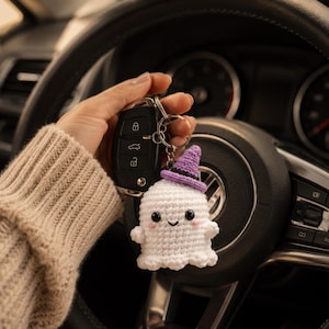 May include: A white crocheted ghost keychain featuring a small purple witch hat with a black band, black bead eyes, a stitched smile, and pink blush accents, attached to a metal keyring with a black car key fob.