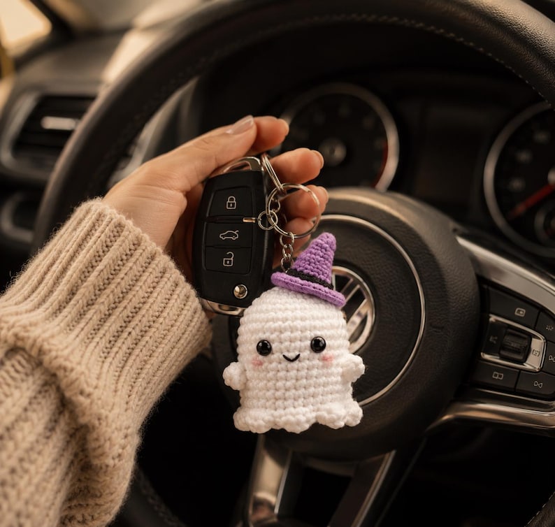 May include: A white crocheted ghost keychain featuring a small purple witch hat with a black band, black bead eyes, a stitched smile, and pink blush accents, attached to a metal keyring with a black car key fob.