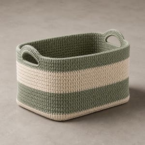 May include: A rectangular storage basket crafted from crocheted yarn, featuring horizontal stripes in sage green and cream. The basket has rounded corners and two integrated handles built into the upper rim on the shorter sides of the structure.