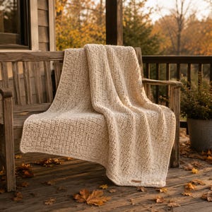 Peut inclure: Un plaid en tricot épais de couleur crème avec un motif de tissage texturé visible. La couverture présente une petite étiquette rectangulaire en similicuir marron fixée dans le coin inférieur, avec une construction en fil épais et doux.