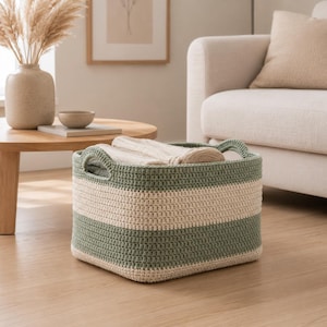 May include: A square-shaped storage basket crafted from thick, crocheted yarn featuring horizontal stripes in sage green and cream. The basket includes two integrated handles on opposite sides and contains a folded, light-colored textile item.