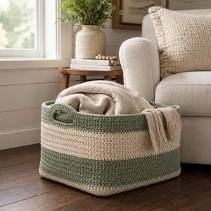 May include: A round, hand-crocheted storage basket featuring alternating horizontal stripes of sage green and cream yarn. The basket has two integrated side handles and contains a folded, textured cream-colored throw blanket with fringe.