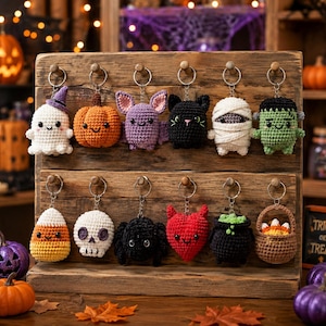 May include: A collection of twelve crocheted Halloween-themed keychains displayed on a wooden rack. The figures include a ghost, pumpkin, bat, black cat, mummy, Frankenstein monster, candy corn, skull, spider, red heart, cauldron, and a basket.