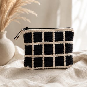 May include: A rectangular crochet pouch featuring a black and cream grid pattern. The accessory is constructed with a textured yarn stitch and includes a black zipper closure along the top edge, complete with a small matching crochet zipper pull.