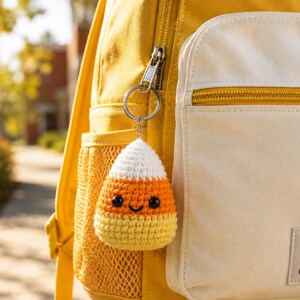May include: A crochet candy corn keychain with a white top, orange middle, and yellow bottom, featuring two black safety eyes and a stitched black smile. The keychain is attached to a silver metal ring hanging from a yellow and white backpack.