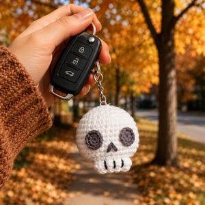 May include: A black plastic car key fob attached to a white crocheted skull keychain. The skull features dark gray circular eye sockets, a small triangular nose, and black stitched vertical lines representing teeth on the lower jaw section.