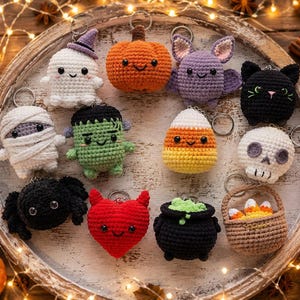 May include: A collection of eleven crocheted Halloween-themed keychains featuring a ghost, pumpkin, bat, black cat, mummy, Frankenstein monster, candy corn, skull, spider, red heart with horns, black cauldron, and a basket of candy corn.