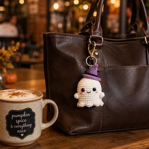 May include: A white crocheted ghost keychain with a purple witch hat, black eyes, and pink cheeks attached to a dark brown leather handbag. Next to the bag is a speckled ceramic mug with the text pumpkin spice & everything nice printed on it.