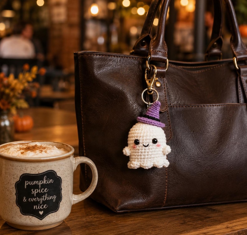 May include: A white crocheted ghost keychain with a purple witch hat, black eyes, and pink cheeks attached to a dark brown leather handbag. Next to the bag is a speckled ceramic mug with the text pumpkin spice & everything nice printed on it.