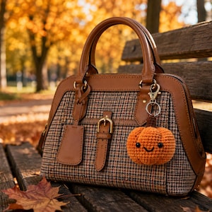 May include: A structured handbag featuring a brown leather trim and a brown, black, and white plaid fabric body. A small, crocheted orange pumpkin charm with a smiling face and black bead eyes is attached to the gold-toned metal hardware.