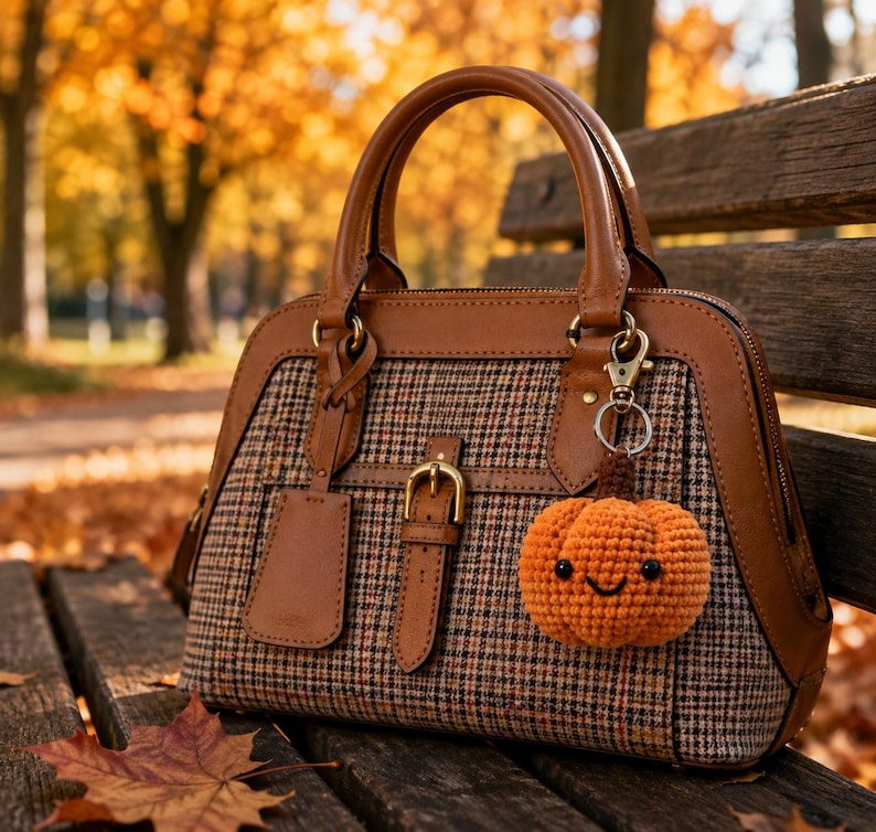 May include: A structured handbag featuring a brown leather trim and a brown, black, and white plaid fabric body. A small, crocheted orange pumpkin charm with a smiling face and black bead eyes is attached to the gold-toned metal hardware.