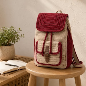 Puede incluir: Una mochila de ganchillo con cuerpo principal en color beige y solapa y paneles laterales en rojo oscuro. La bolsa incluye un bolsillo frontal con una correa de piel sintética marrón y cierre de hebilla, cordones rojos y dos correas de ganchillo.