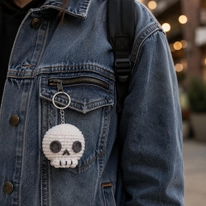 May include: A white crocheted skull keychain featuring dark gray circular eye sockets and a small triangular nose. The skull has a stitched mouth line and is attached to a silver-toned metal split ring and a short chain, hanging from a denim jacket.