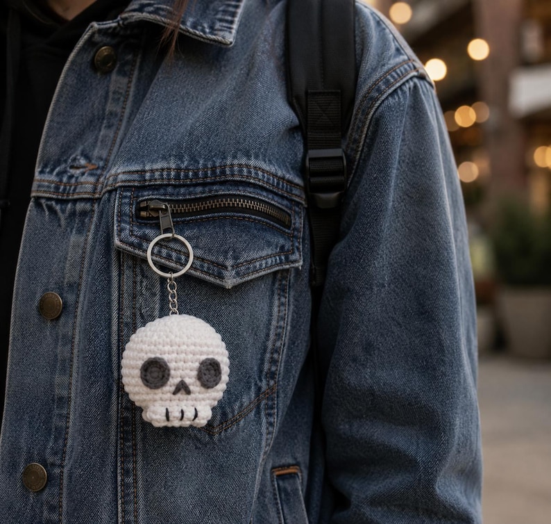 May include: A white crocheted skull keychain featuring dark gray circular eye sockets and a small triangular nose. The skull has a stitched mouth line and is attached to a silver-toned metal split ring and a short chain, hanging from a denim jacket.