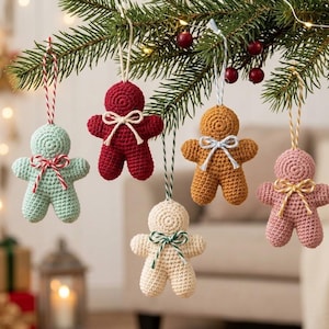 May include: Five crocheted gingerbread-style ornaments in light green, dark red, mustard yellow, cream, and dusty pink. Each figure features a small tied bow at the neck and a matching hanging string, suspended from a green pine branch.