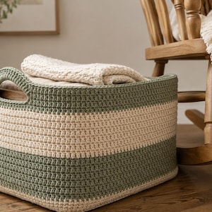 May include: A rectangular crochet storage basket featuring alternating horizontal stripes of sage green and cream yarn. The basket has integrated handles on the sides and contains a folded cream-colored textile item inside the main compartment.