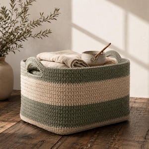 May include: A rectangular woven storage basket featuring horizontal stripes in sage green and cream. The basket has two integrated handles on the sides and is filled with folded beige fabric and a small ceramic bowl with a wooden spoon inside.