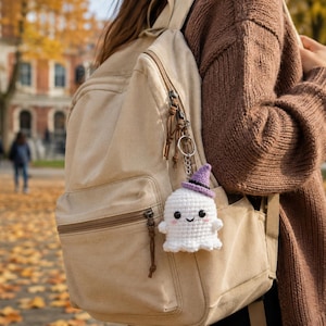 May include: A white crocheted ghost keychain featuring a small purple witch hat, black bead eyes, and pink stitched cheeks, attached to a silver metal keyring hanging from the side zipper of a beige canvas backpack.