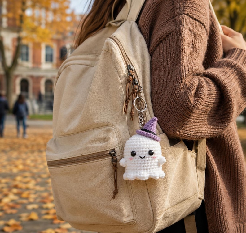 May include: A white crocheted ghost keychain featuring a small purple witch hat, black bead eyes, and pink stitched cheeks, attached to a silver metal keyring hanging from the side zipper of a beige canvas backpack.